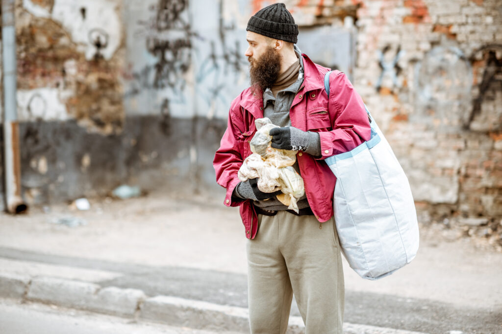 Fentanyl Addiction Signs: Homeless depressed beggar standing with bags on the street on the ragged wall background